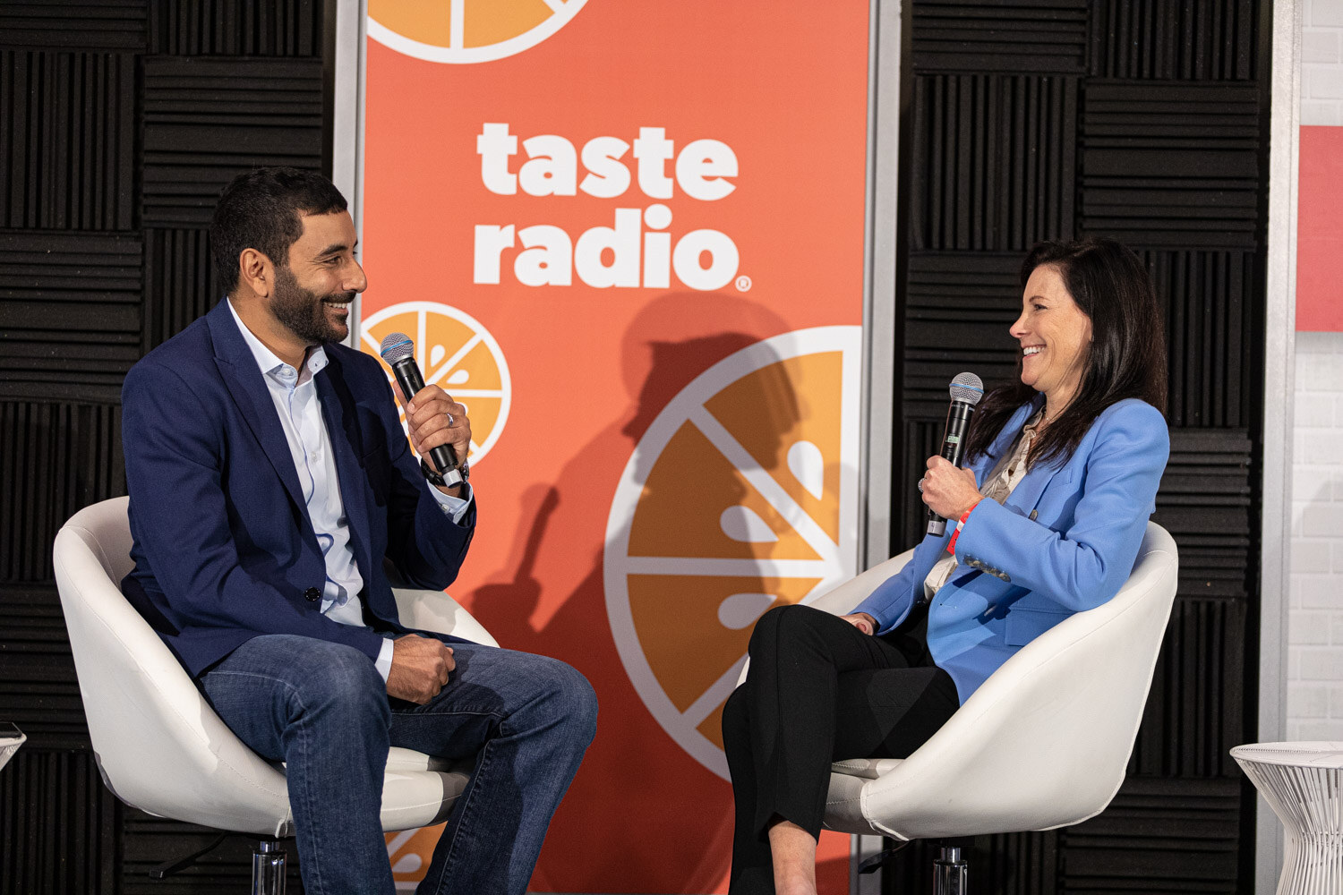 A man in a navy blazer and a woman in a light blue blazer, both smiling and holding microphones, sit in white chairs in front of a Taste Radio banner.
