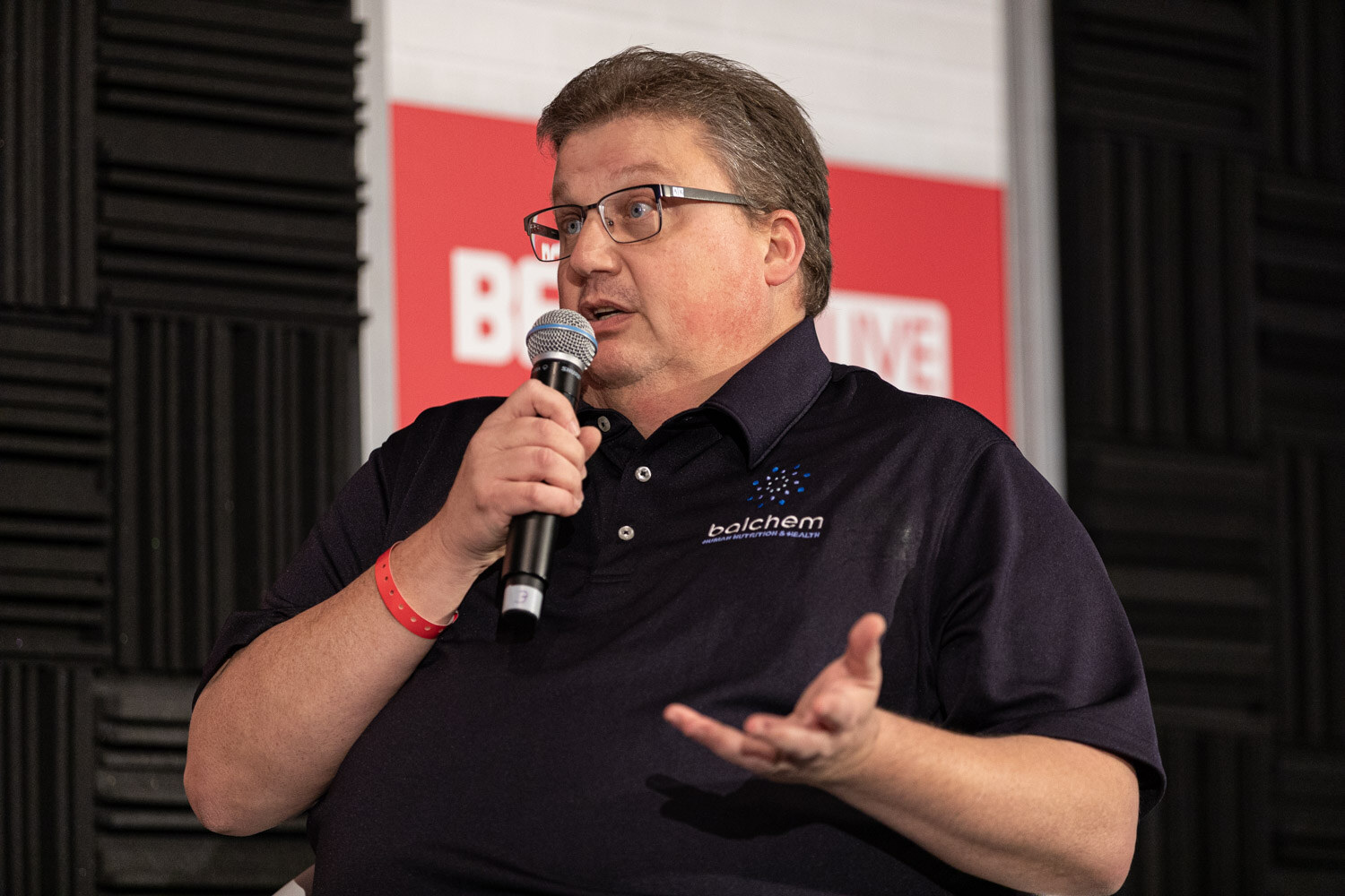 A man in glasses and a navy Balchem polo holds a microphone and gestures with his other hand in front of a BevNET Live sign.