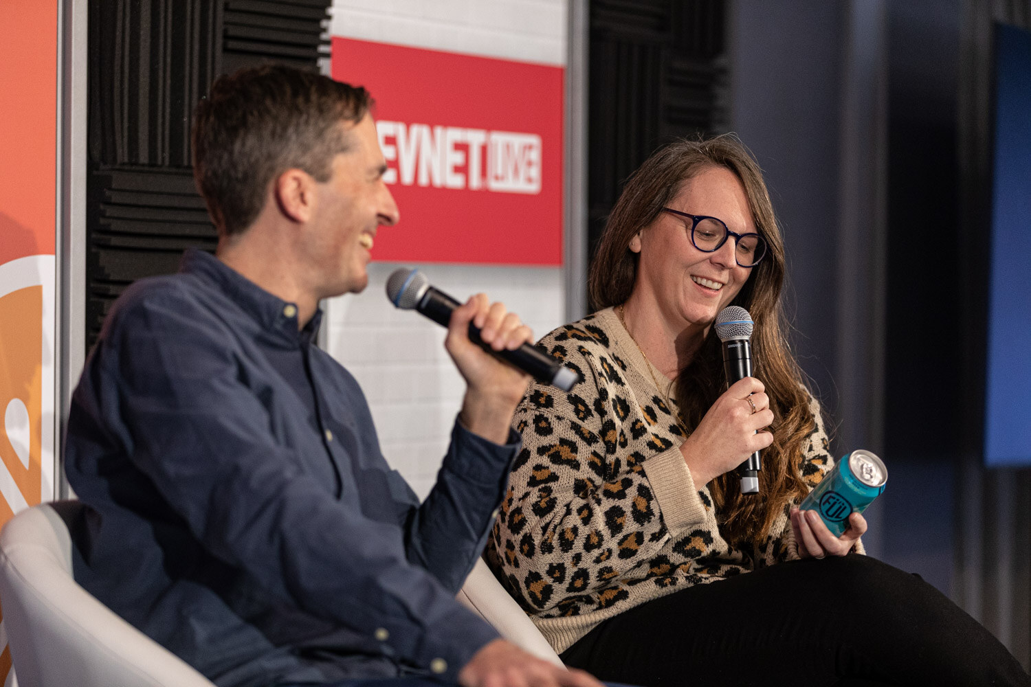 A man in a navy shirt talks into a microphone while a woman in a leopard-print sweater holds a teal can and laughs.