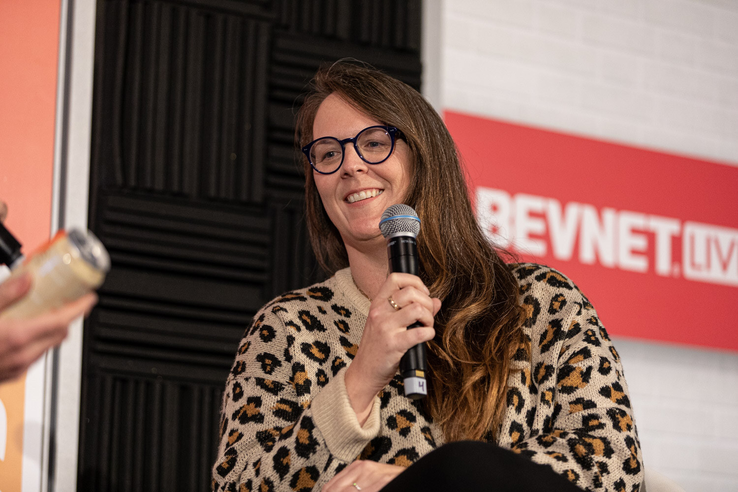 A woman in glasses and a leopard-print sweater holds a microphone and smiles in front of a BevNET Live sign.