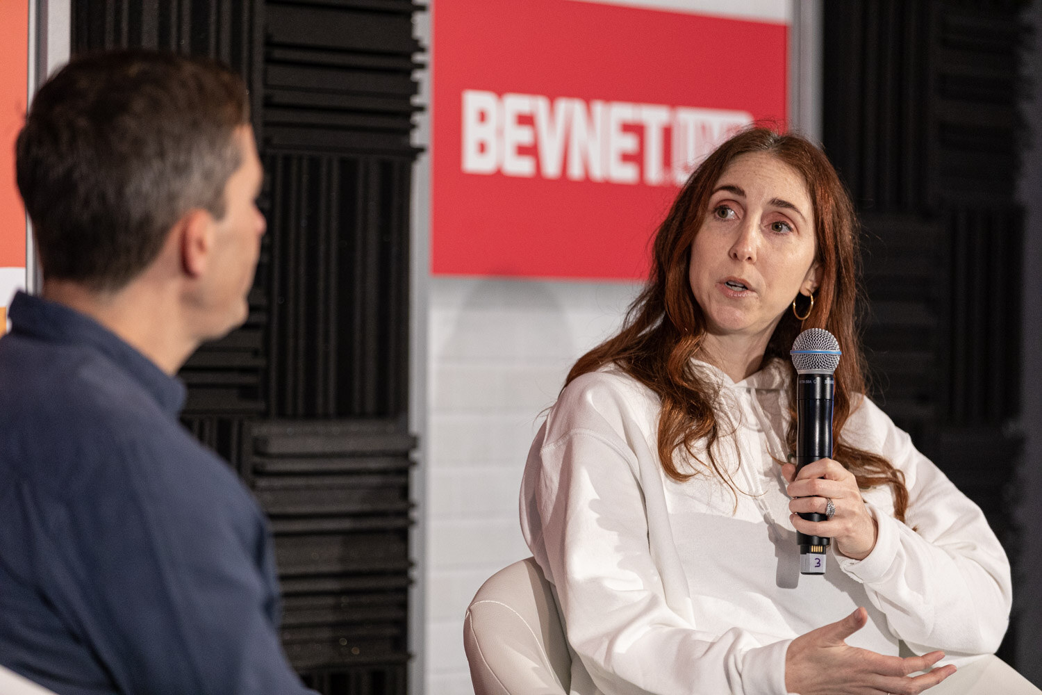 A woman with long auburn hair in a white sweatshirt holds a microphone and speaks while a man in a navy shirt sits with his back to the camera.