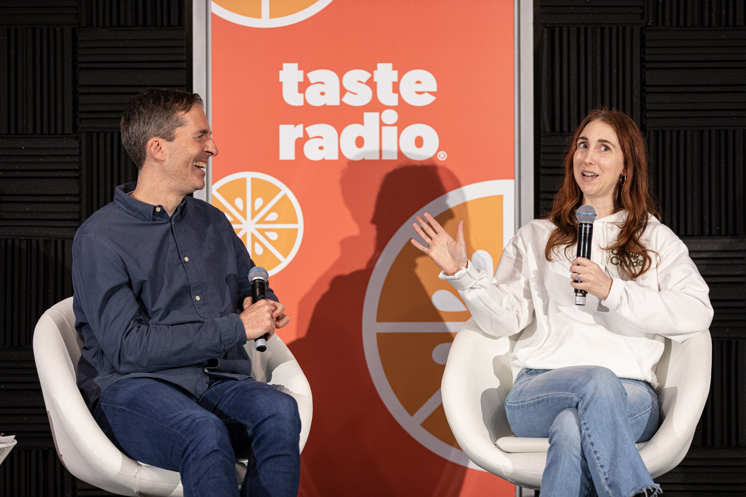 A man in a navy shirt laughs while a woman in a white hoodie and jeans gestures with a microphone in white chairs in front of a Taste Radio banner.