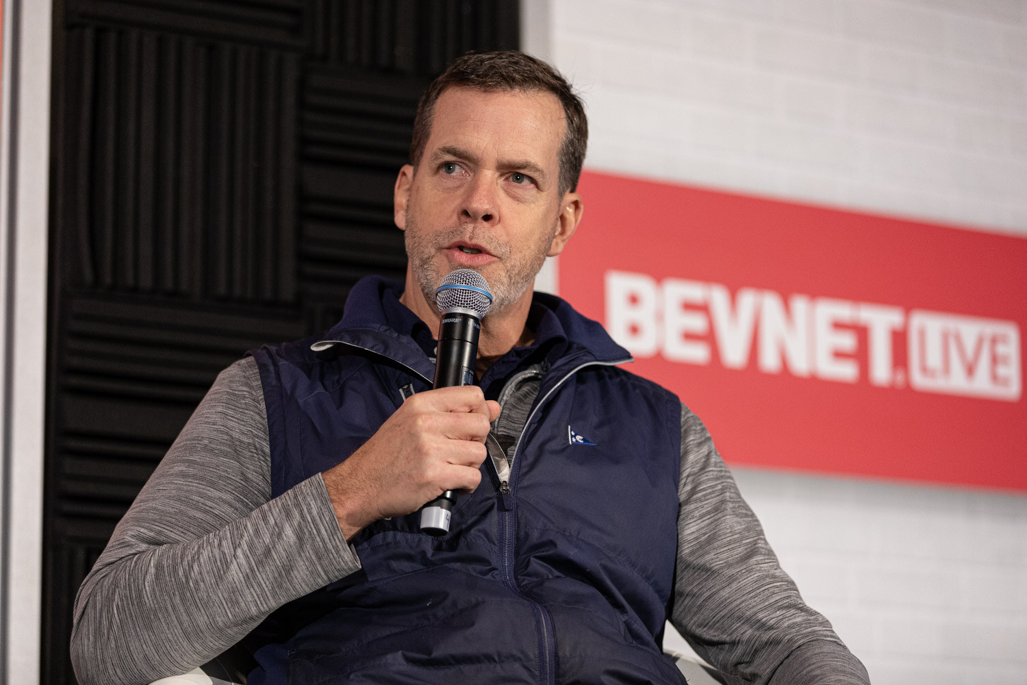 A man in a navy vest and gray long-sleeve shirt holds a microphone in front of a BevNET Live sign.