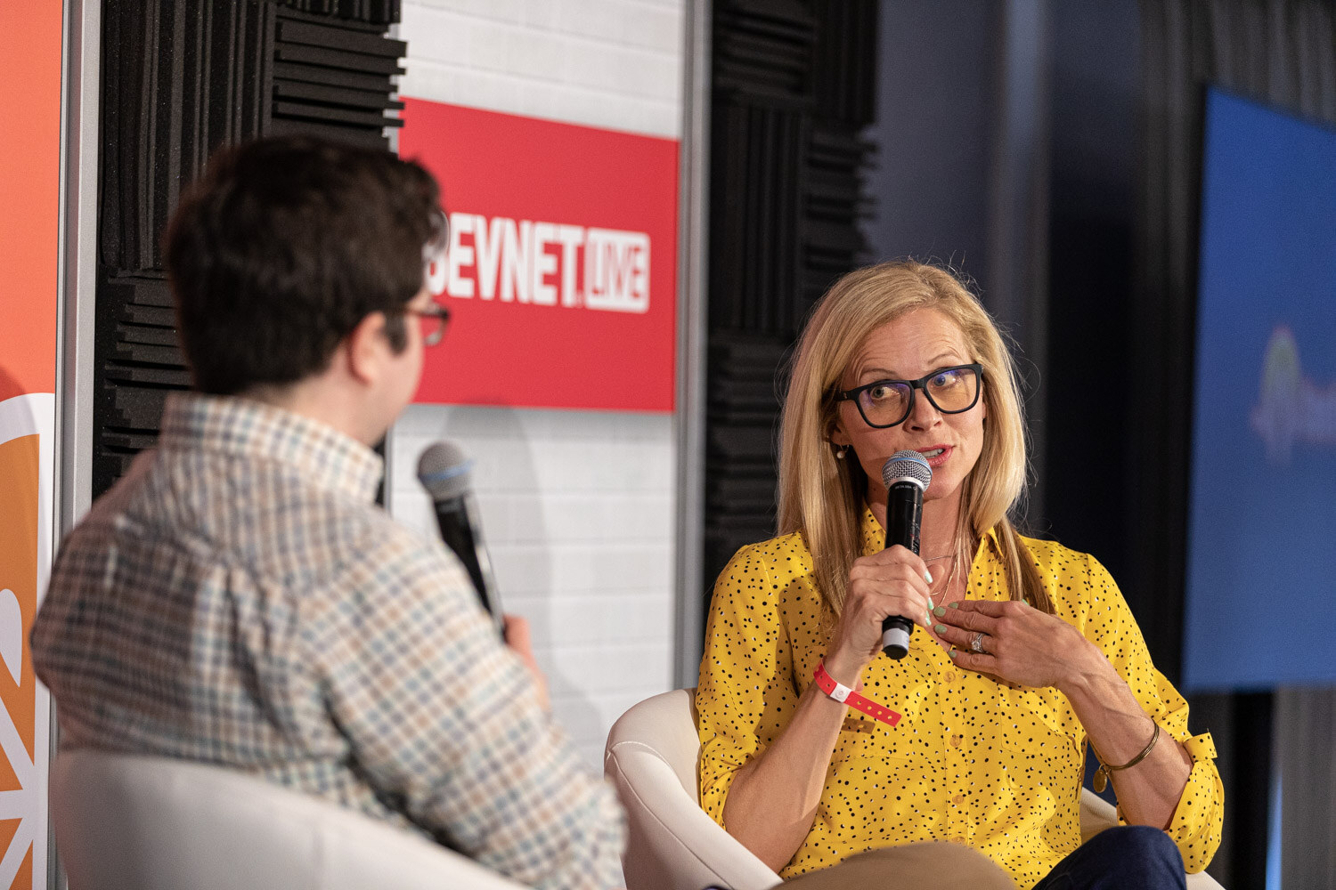 A blond woman in glasses and a yellow blouse holds a microphone with one hand and presses the other to her chest.