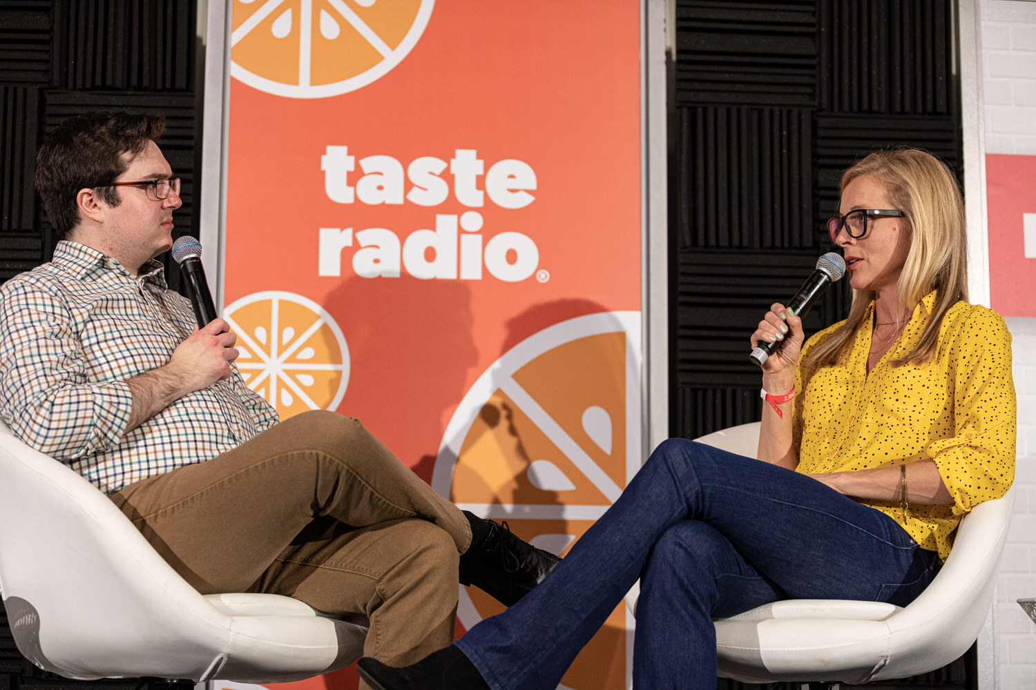A man in a checked shirt and a woman in a yellow blouse and jeans sit in white chairs in front of a Taste Radio banner.
