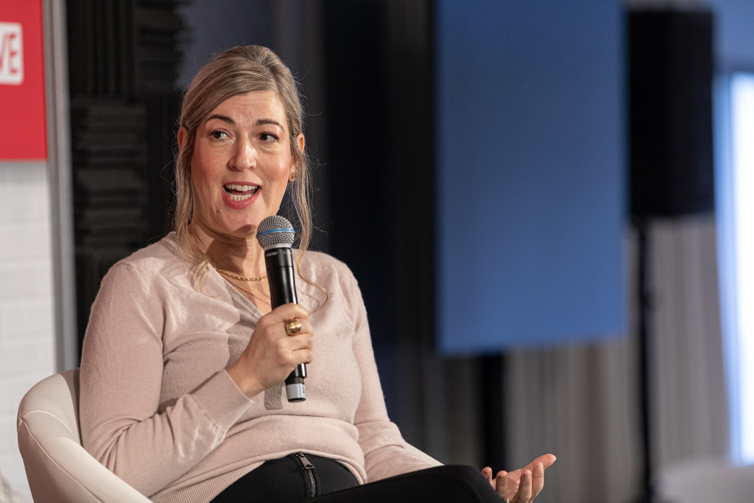A woman with light hair in a pink sweater holds a microphone and gestures with an open hand.