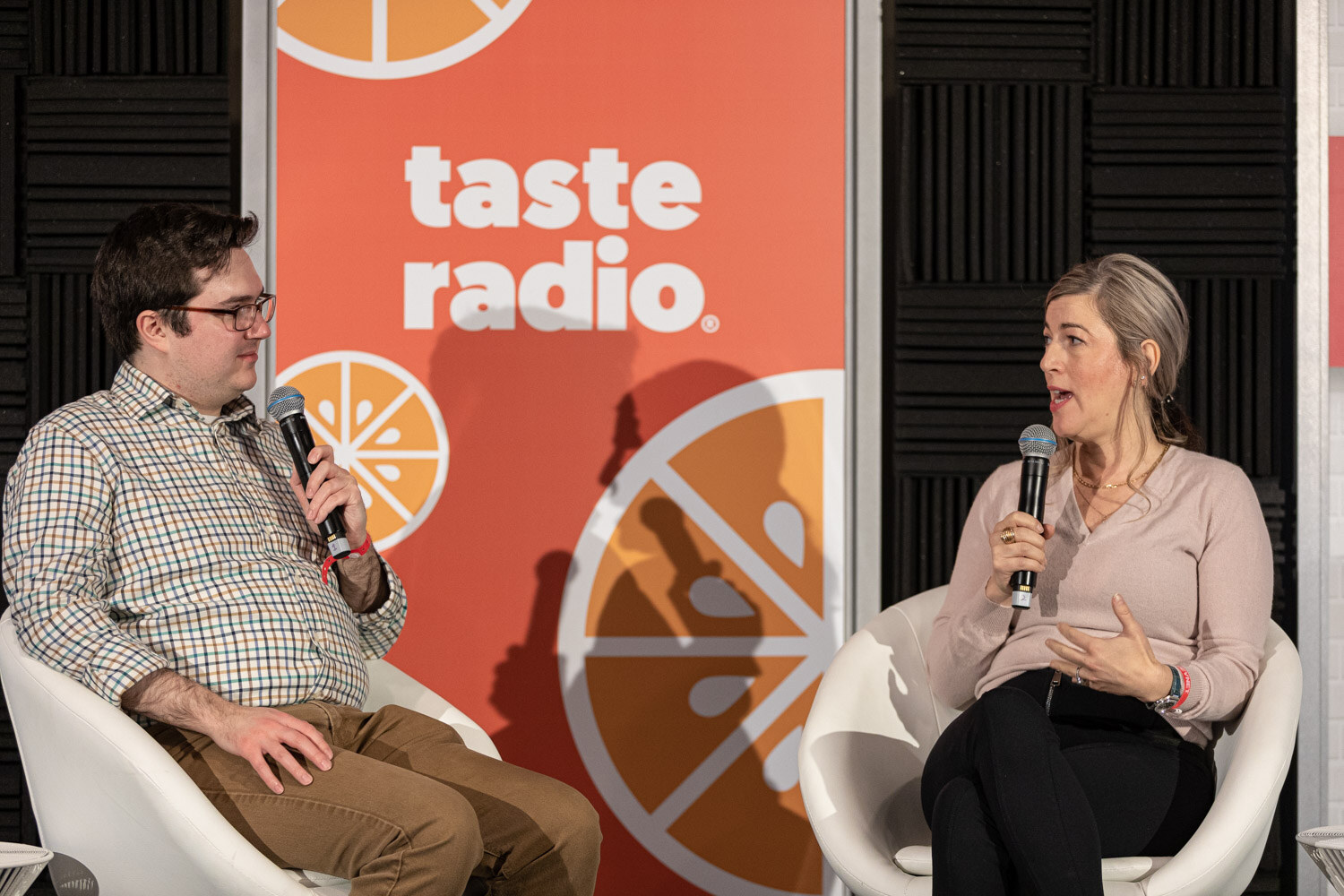 A man in a checked shirt holding a microphone and a woman in a pink sweater and black pants sit in white chairs in front of a Taste Radio banner.