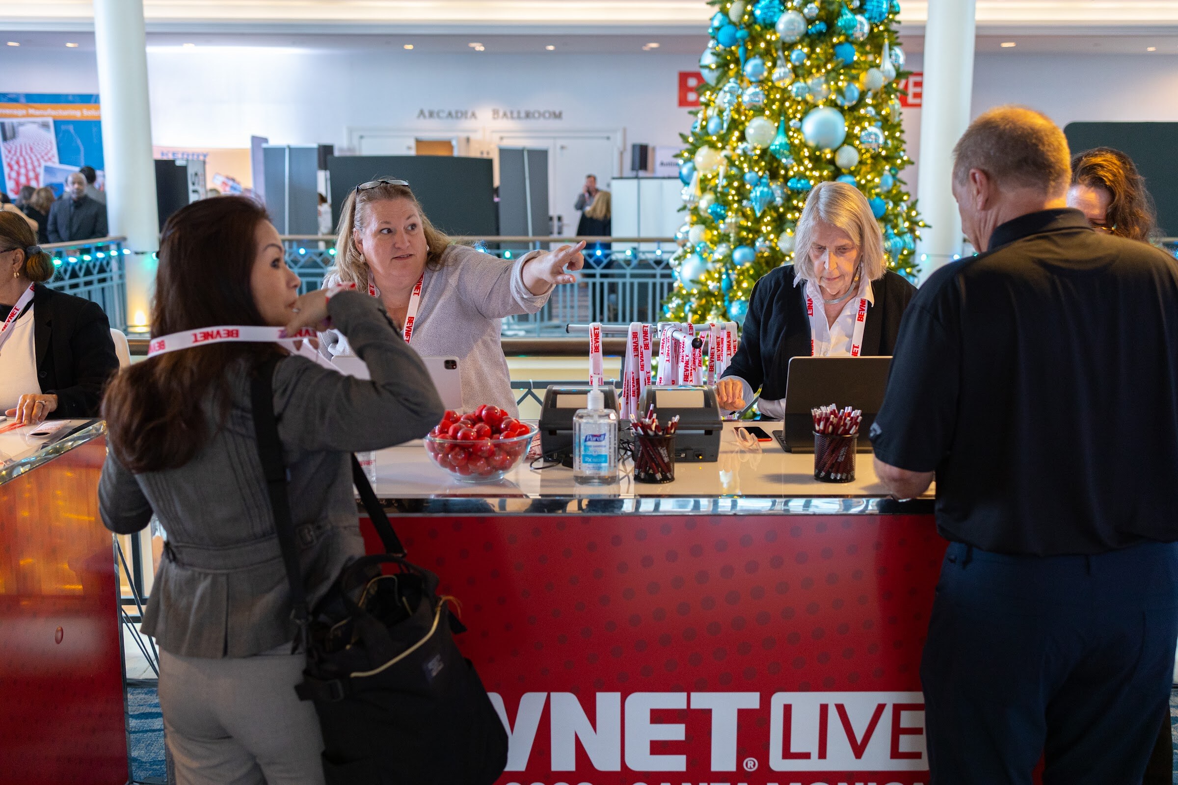 Registration staff at a red BevNET Live desk hand out lanyards and point as two attendees stand at the counter, with a lit holiday tree behind them.
