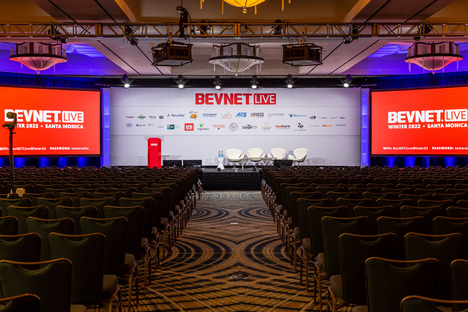 A wide view of rows of teal chairs facing a stage with white swan chairs, a red podium and a BevNET Live wall of sponsor logos, flanked by screens.