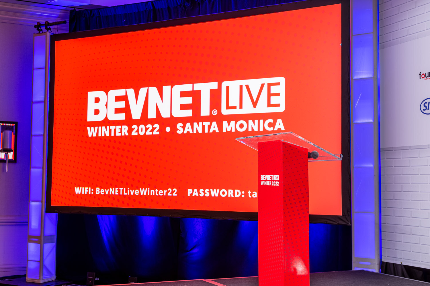 A large red screen reading BevNET Live Winter 2022 Santa Monica with Wi-Fi details beside a red podium on a blue-lit stage.