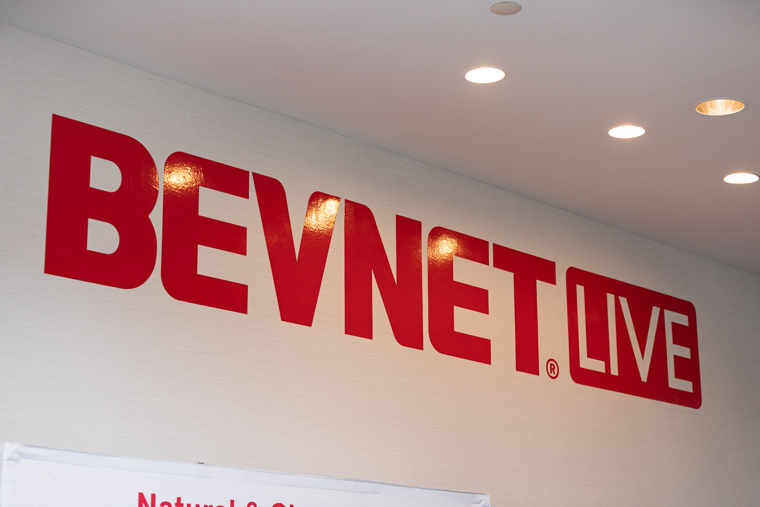 Large red BevNET Live lettering on a white wall with recessed ceiling lights above.