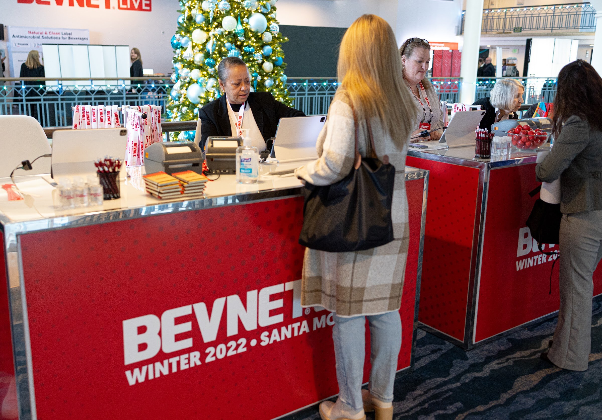 A woman stands at a red registration desk branded BevNET Winter 2022 Santa Monica while staff behind it help her, with a decorated Christmas tree in the background.