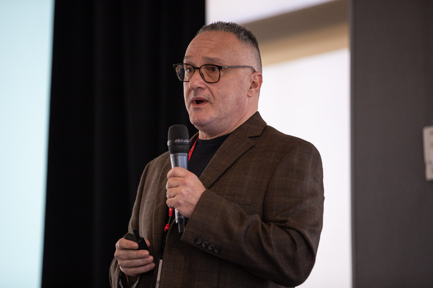 A man in a brown blazer holds a microphone and speaks to a room, with a dark curtain behind him.