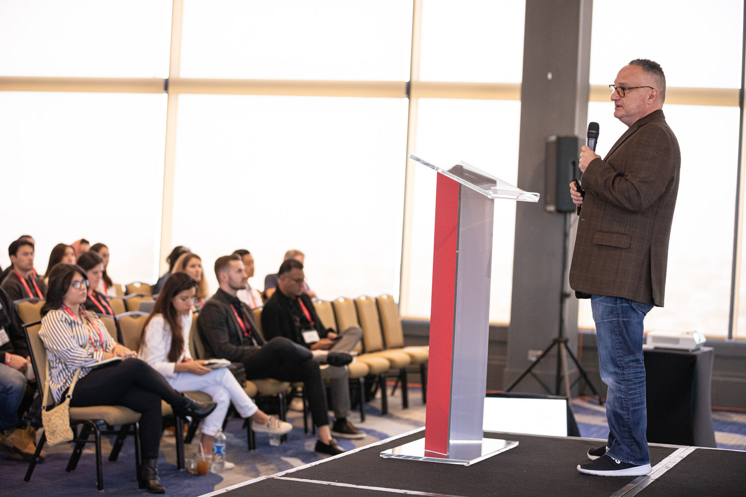 A man in a blazer stands beside a red lectern while attendees sit in chairs near floor-to-ceiling windows.