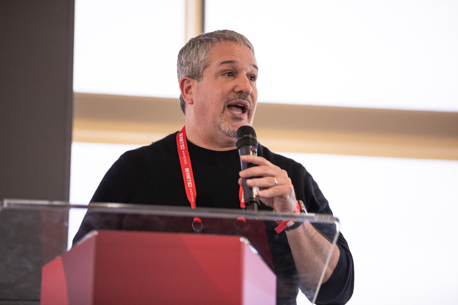 A man with gray hair in a black shirt and red lanyard speaks into a microphone behind a clear lectern.