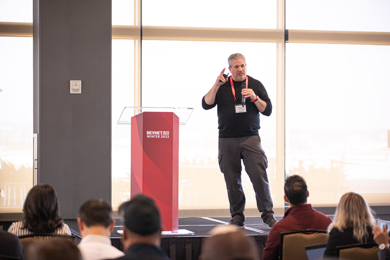A man in a dark shirt gestures on a low stage beside a BevNET Live lectern while attendees watch from the foreground.