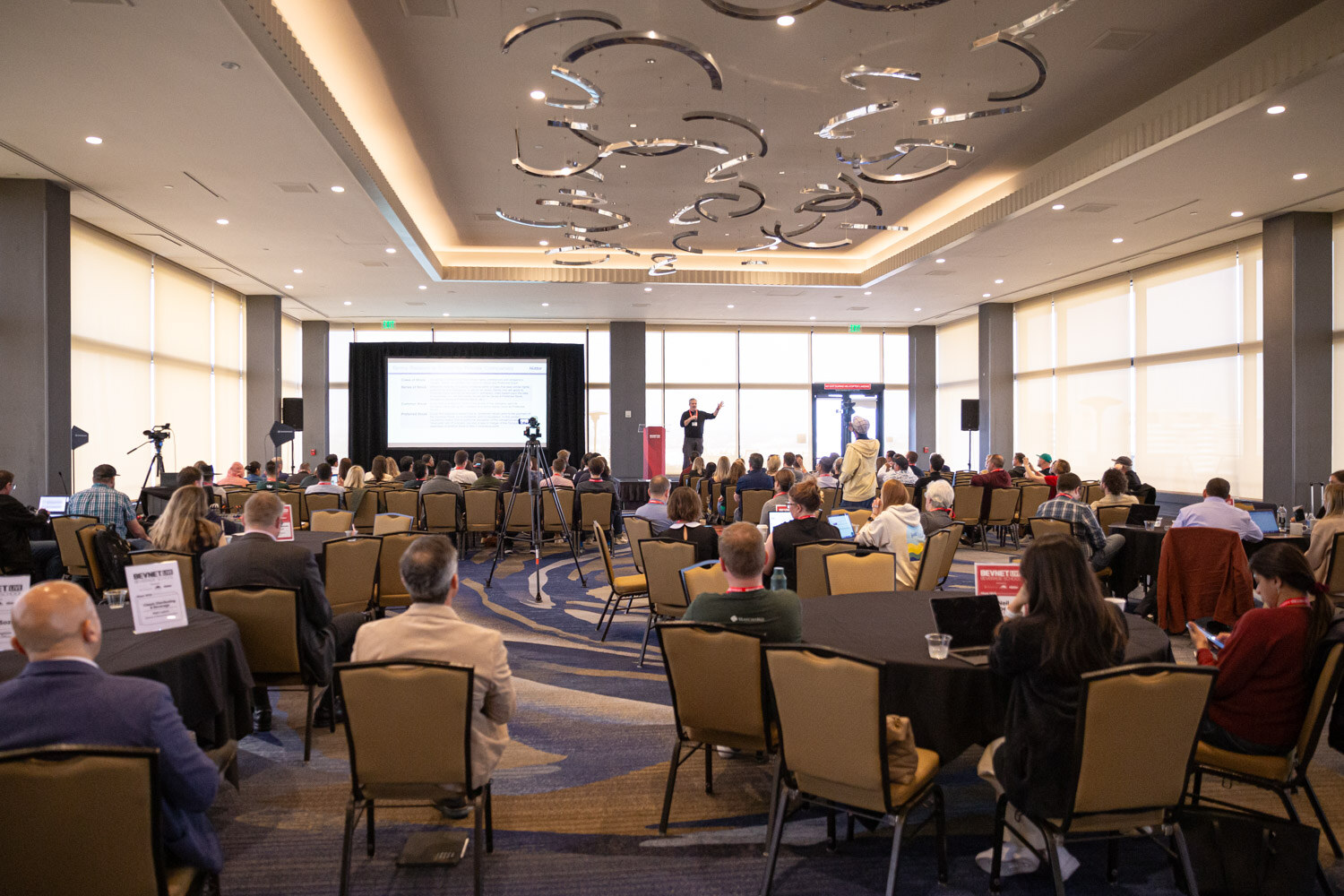 A wide view of a ballroom with round tables, attendees seated and a presentation screen at the far end.