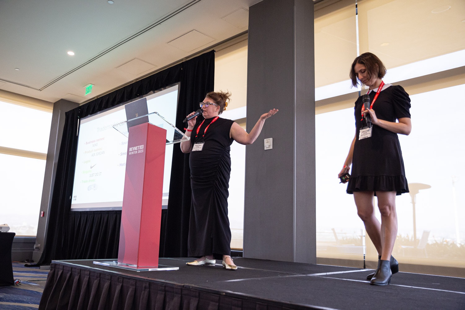 Two women stand on a low stage beside a red lectern, one speaking with a microphone while gesturing.