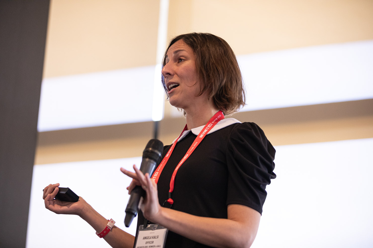 A woman in a black dress with a white collar holds a microphone and a clicker while speaking.