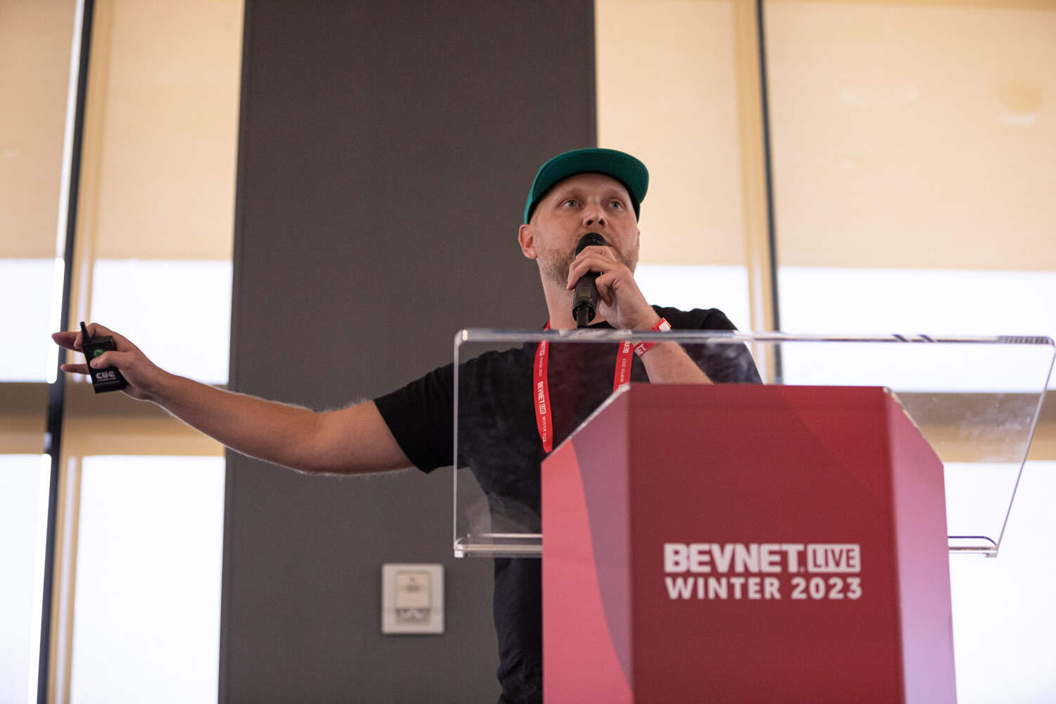 A man in a green cap and black shirt gestures at a lectern marked BevNET Live Winter 2023.
