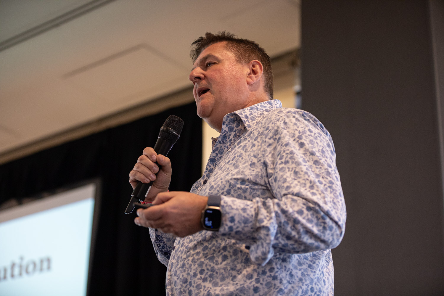 A man in a blue patterned shirt and smart watch holds a microphone and speaks.