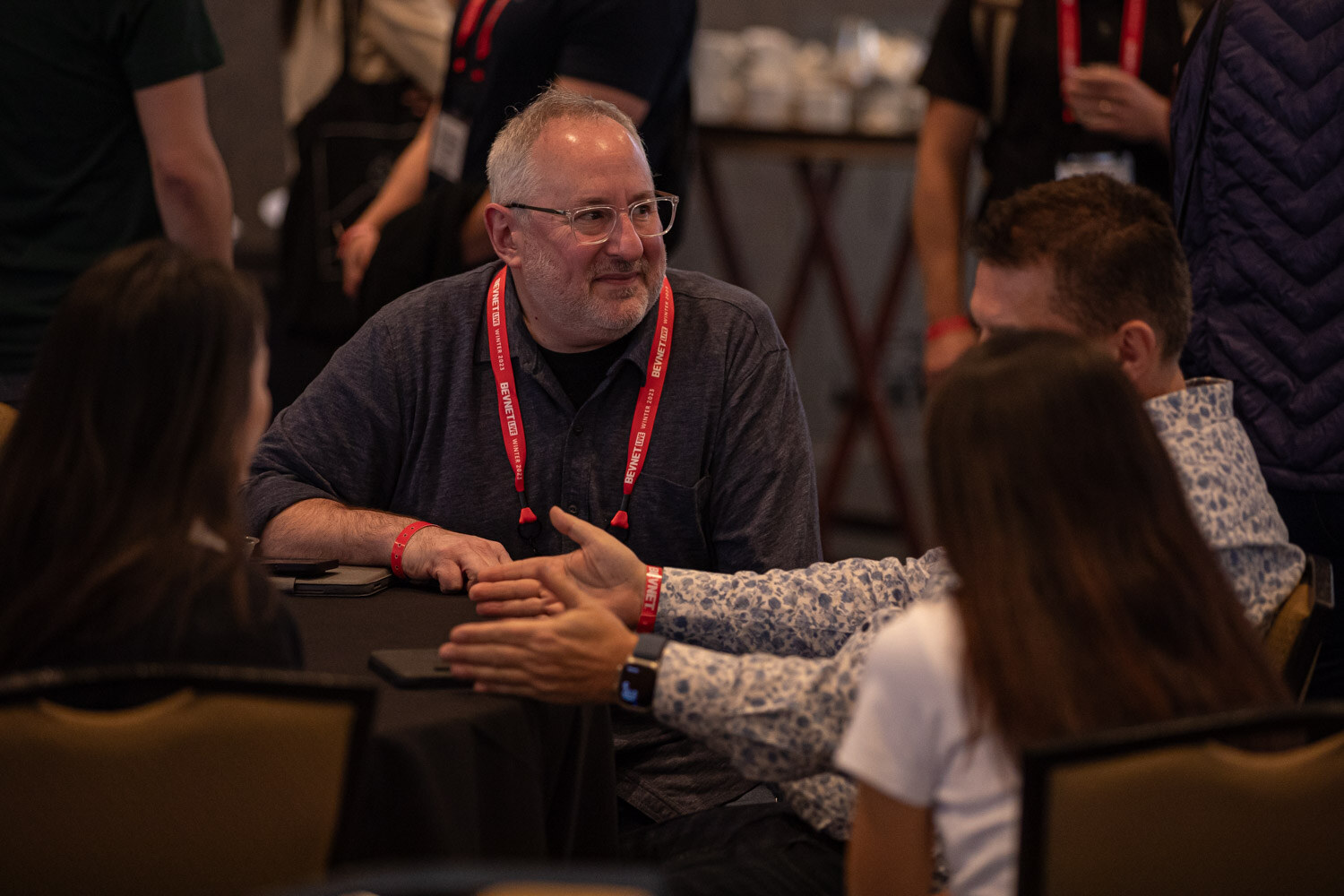 Several attendees in red lanyards sit around a table and shake hands in conversation.