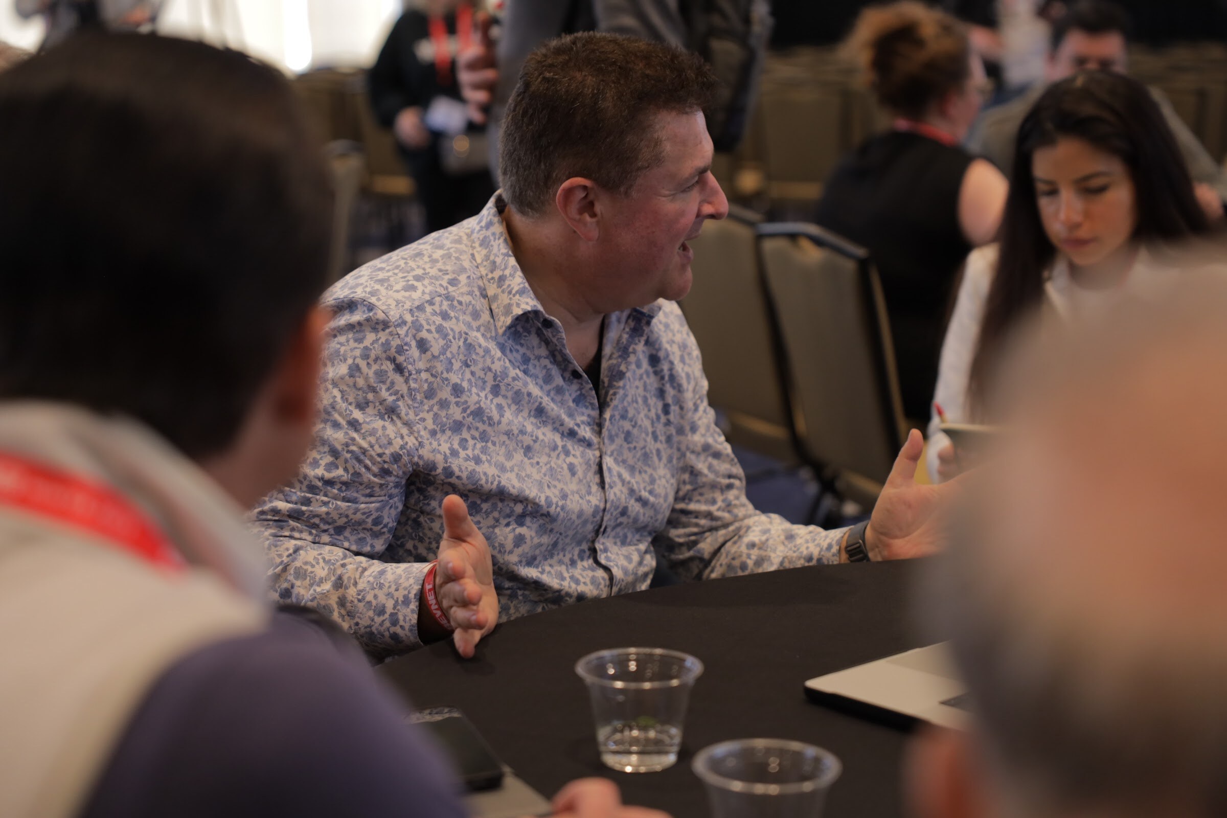 A man in a patterned shirt gestures while talking at a crowded table in a ballroom.