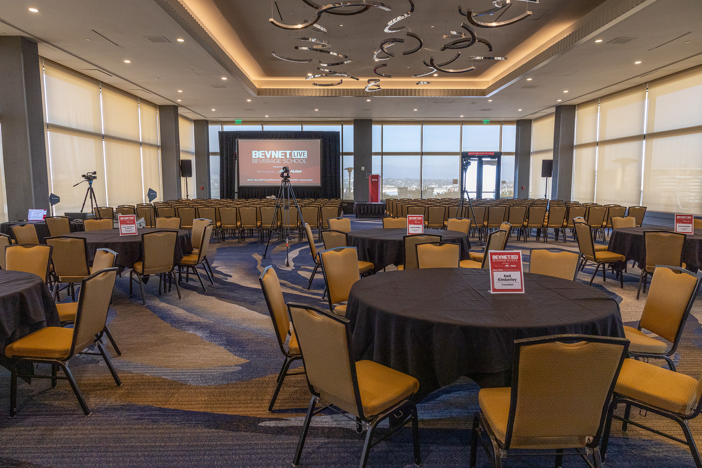 A wide view of a large ballroom with round tables and chairs set up, with a presentation screen far in the background.