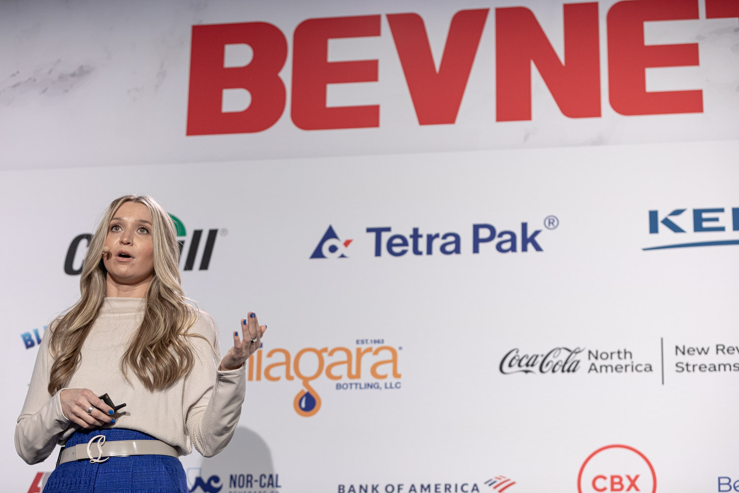 A woman with long blond hair in a cream sweater and blue pants speaks to the room.