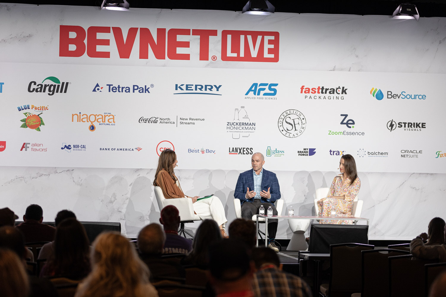 Three people sit on a stage in front of a BevNET Live sponsor wall with an audience in the foreground.