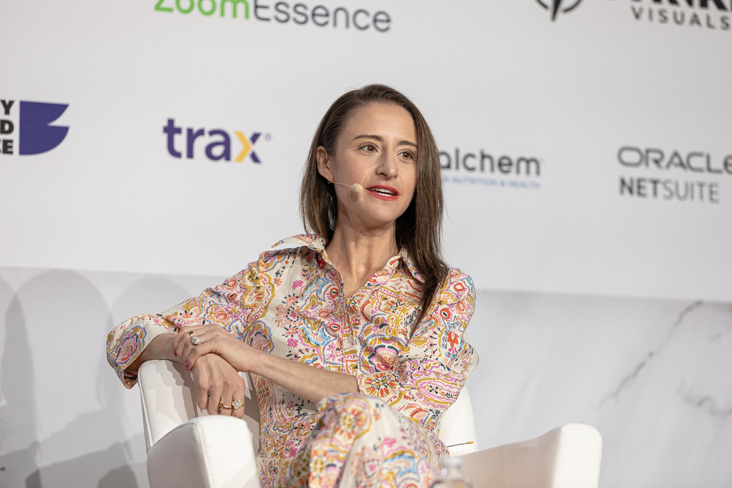 A woman in a floral dress sits on stage in front of a white sponsor backdrop.