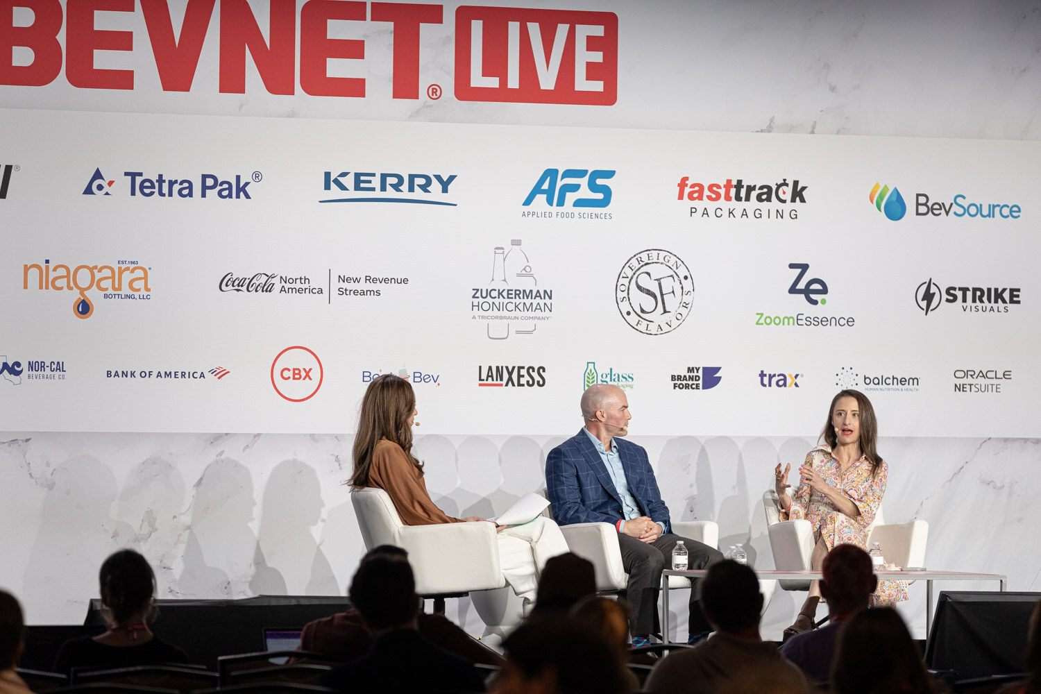 Three people sit on stage; a large screen to the right shows one panelist.