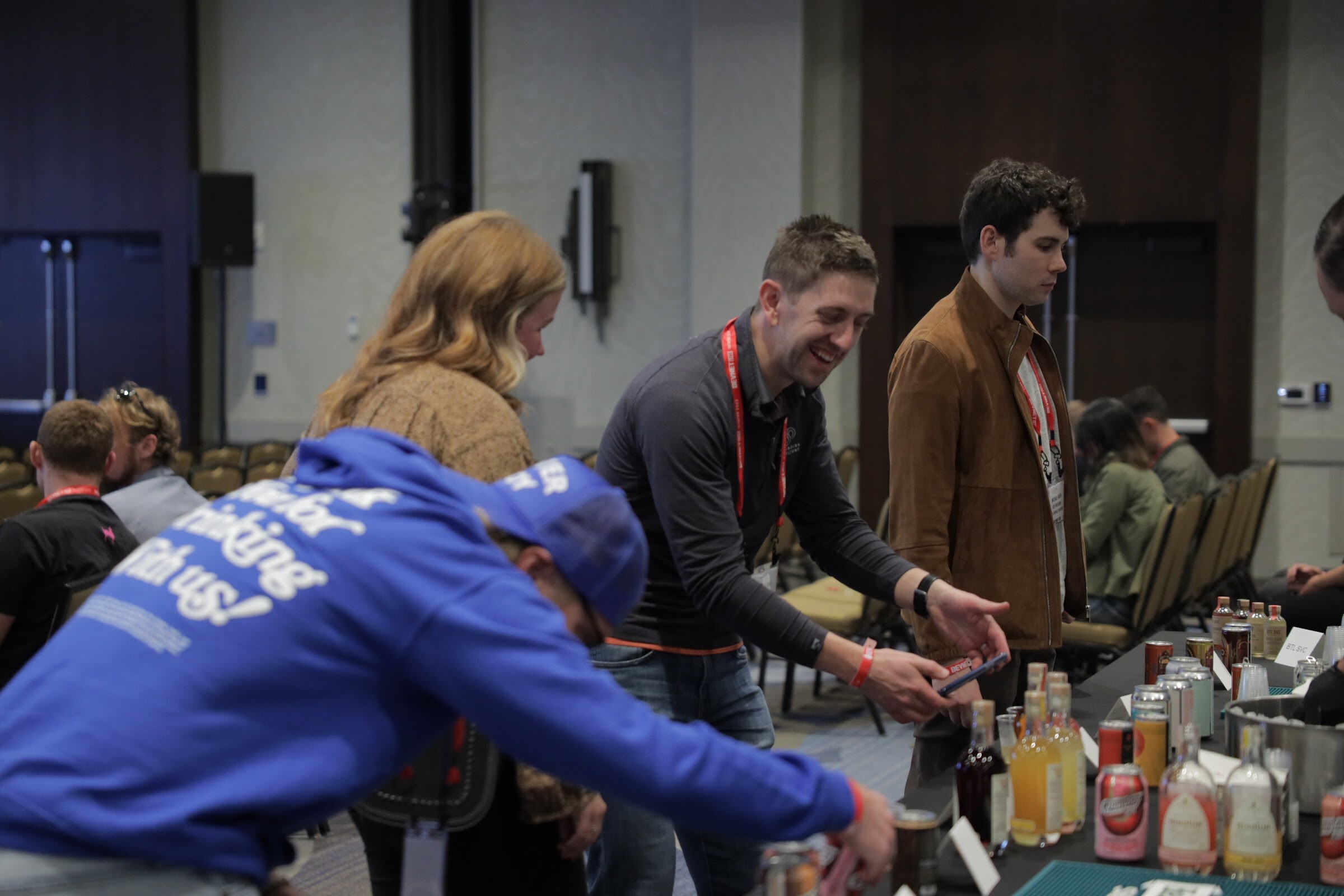 A man leans over a table of bottles while attendees stand beside him.
