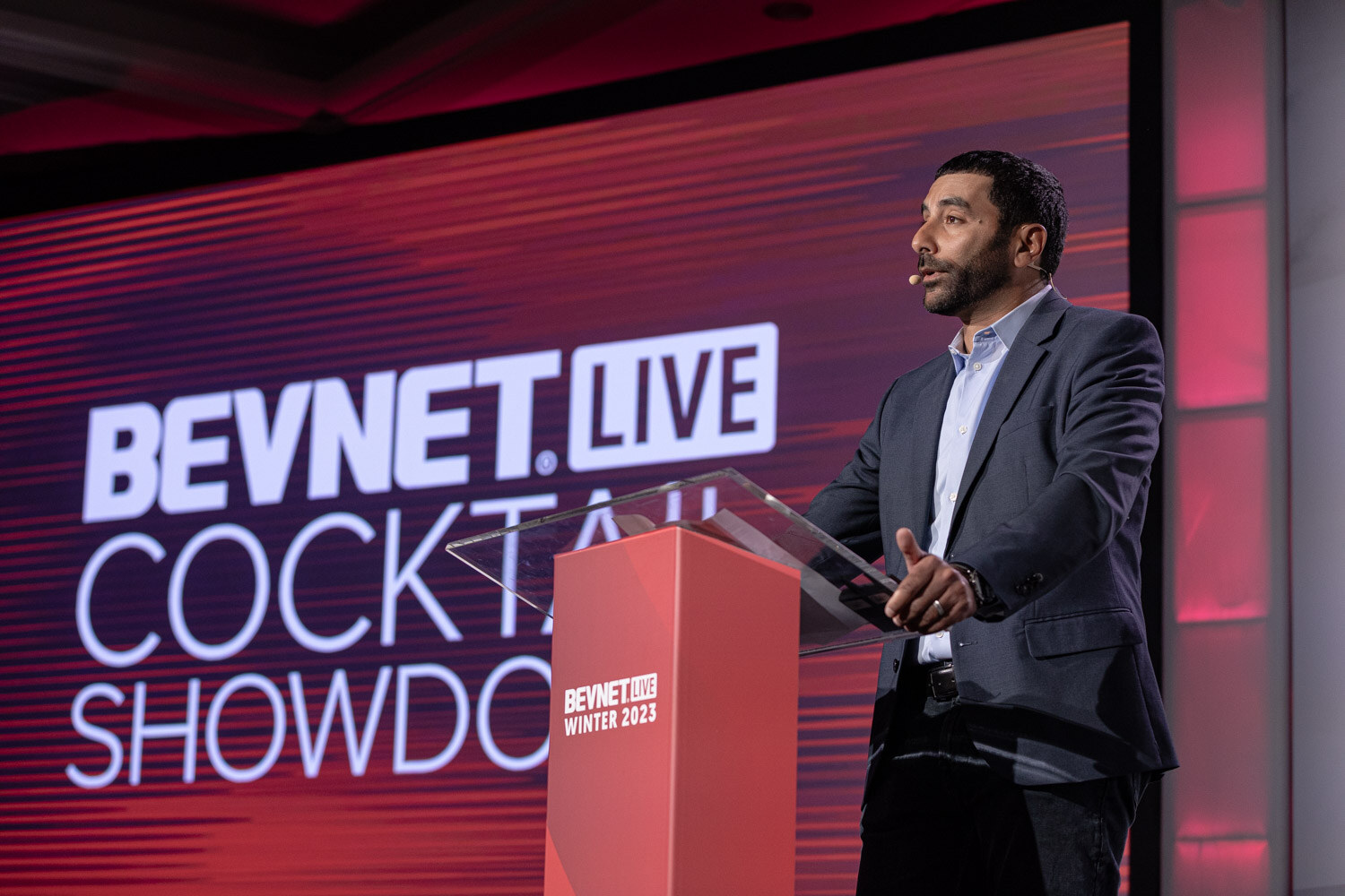 A man in a gray jacket stands at a lectern in front of a red Cocktail Showdown screen.