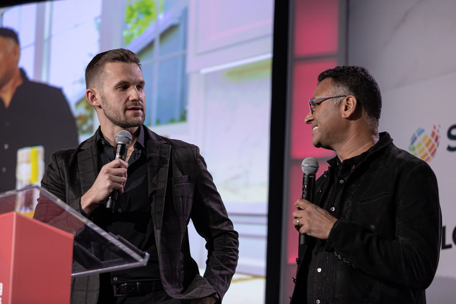 A man in a dark jacket and a man in a black shirt speak into microphones at a red lectern in front of a screen.