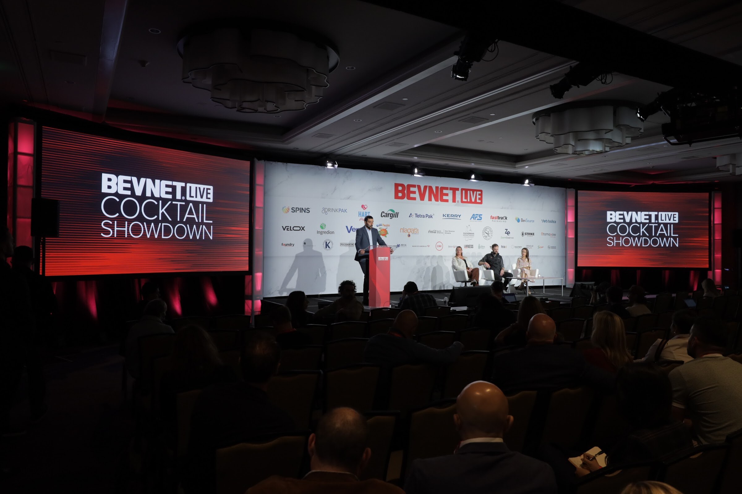 A wide view of a dim ballroom with a red Cocktail Showdown screen and a speaker on stage.