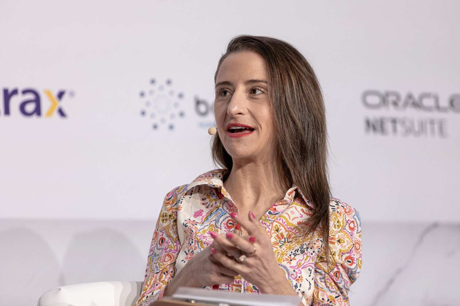 A woman in a floral blouse speaks while seated in front of a sponsor wall.