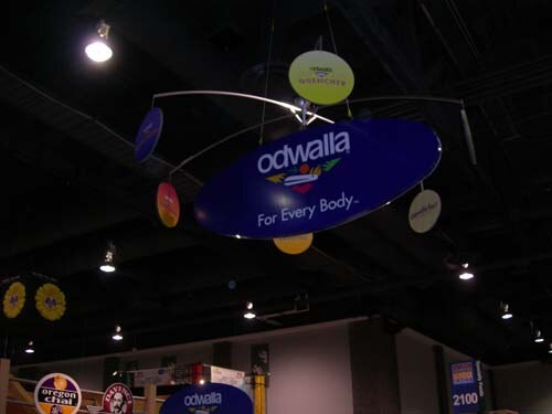 Dark exhibit hall ceiling with a hanging mobile of oval signs, the largest reading Odwalla, For Every Body. An Oregon Chai sign and another Odwalla sign are visible at the bottom.