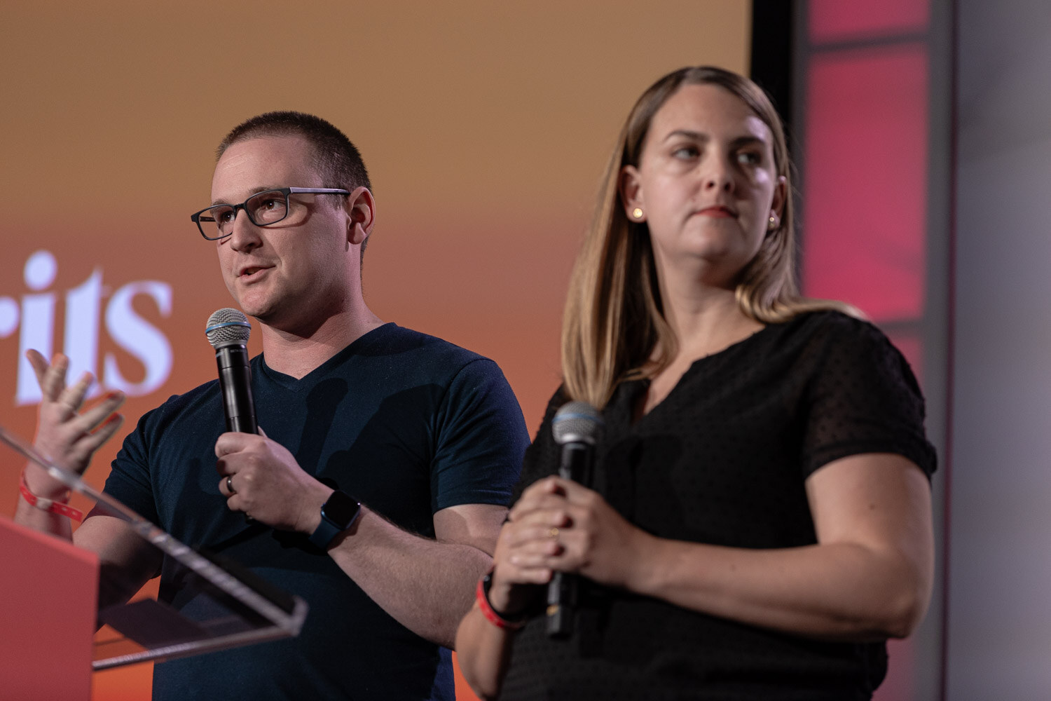 A man in glasses and a woman in a black shirt hold microphones in front of an orange slide.