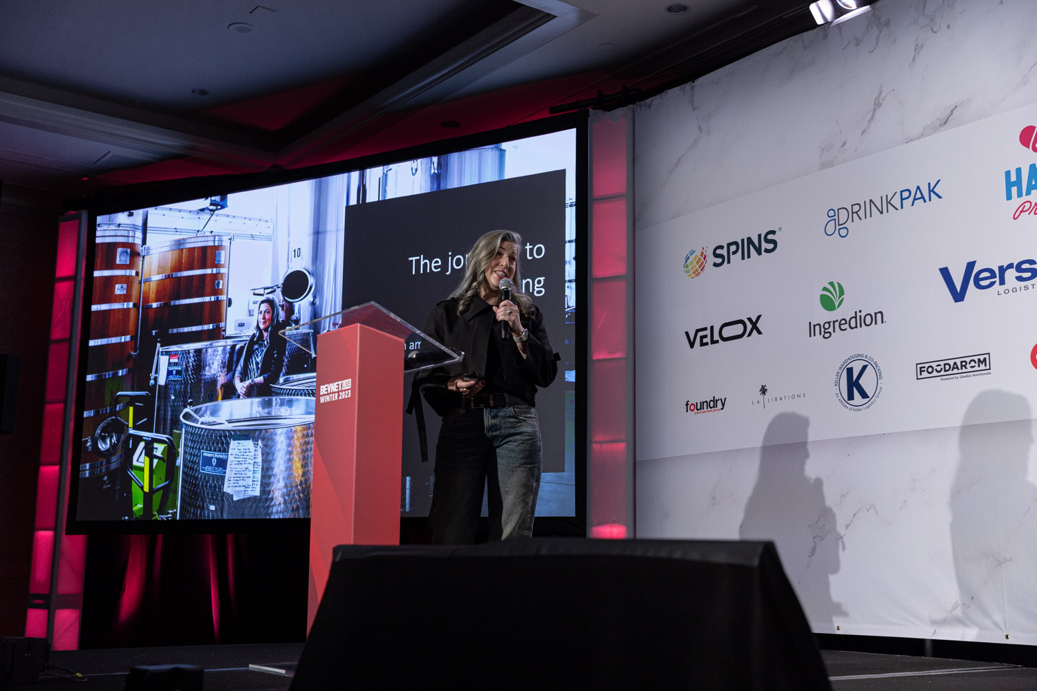 A woman stands at a red lectern on a dark stage as a distillery video plays on a screen.
