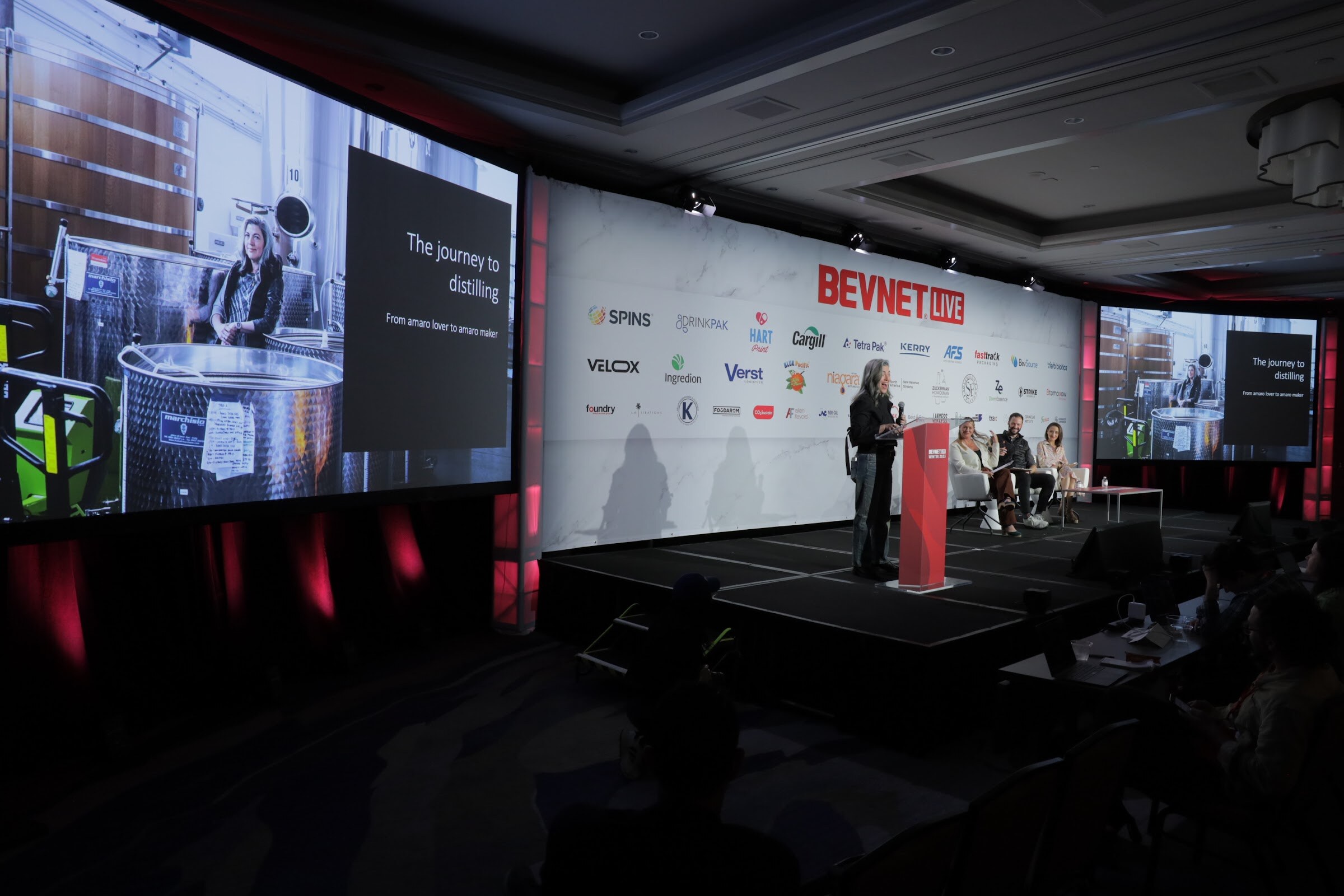 A large video screen shows a distillery while a woman stands at a lectern and judges sit on the stage.