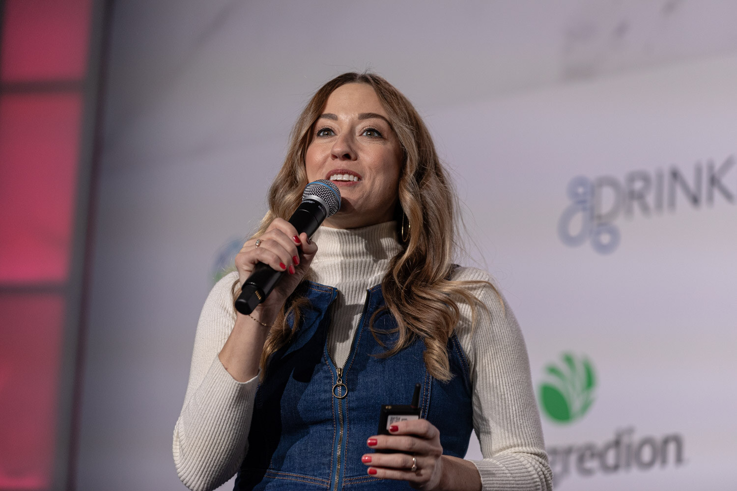 A woman with long wavy hair in a white sweater and denim vest holds a microphone.