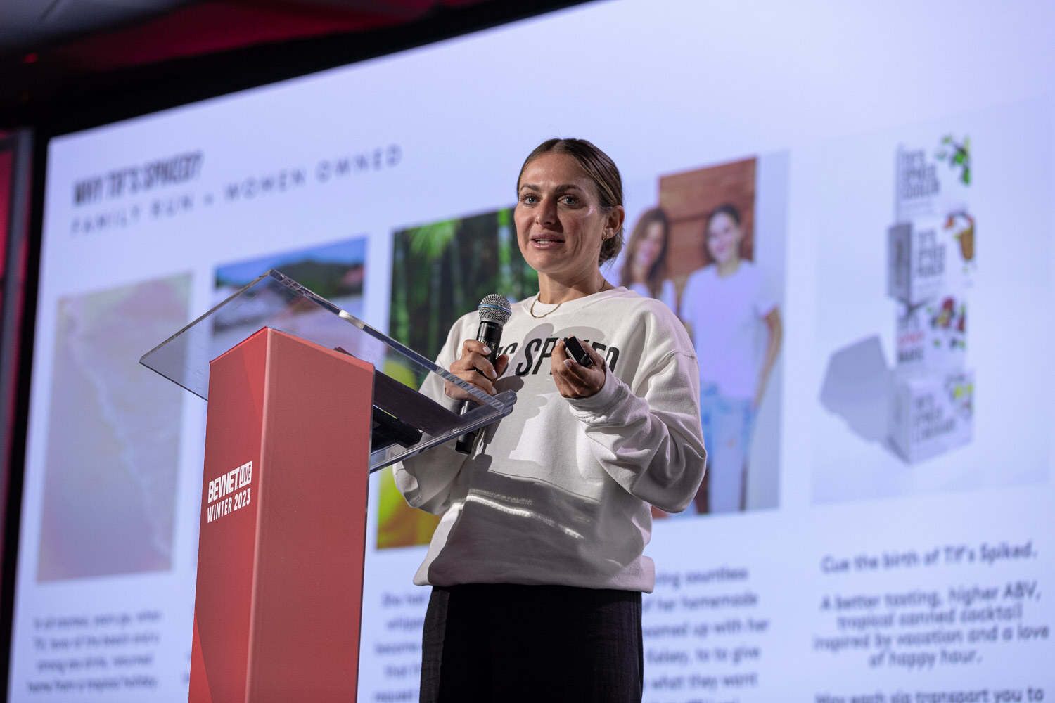 A woman in a gray sweatshirt stands at a lectern in front of a slide describing a family-run, women-owned brand.