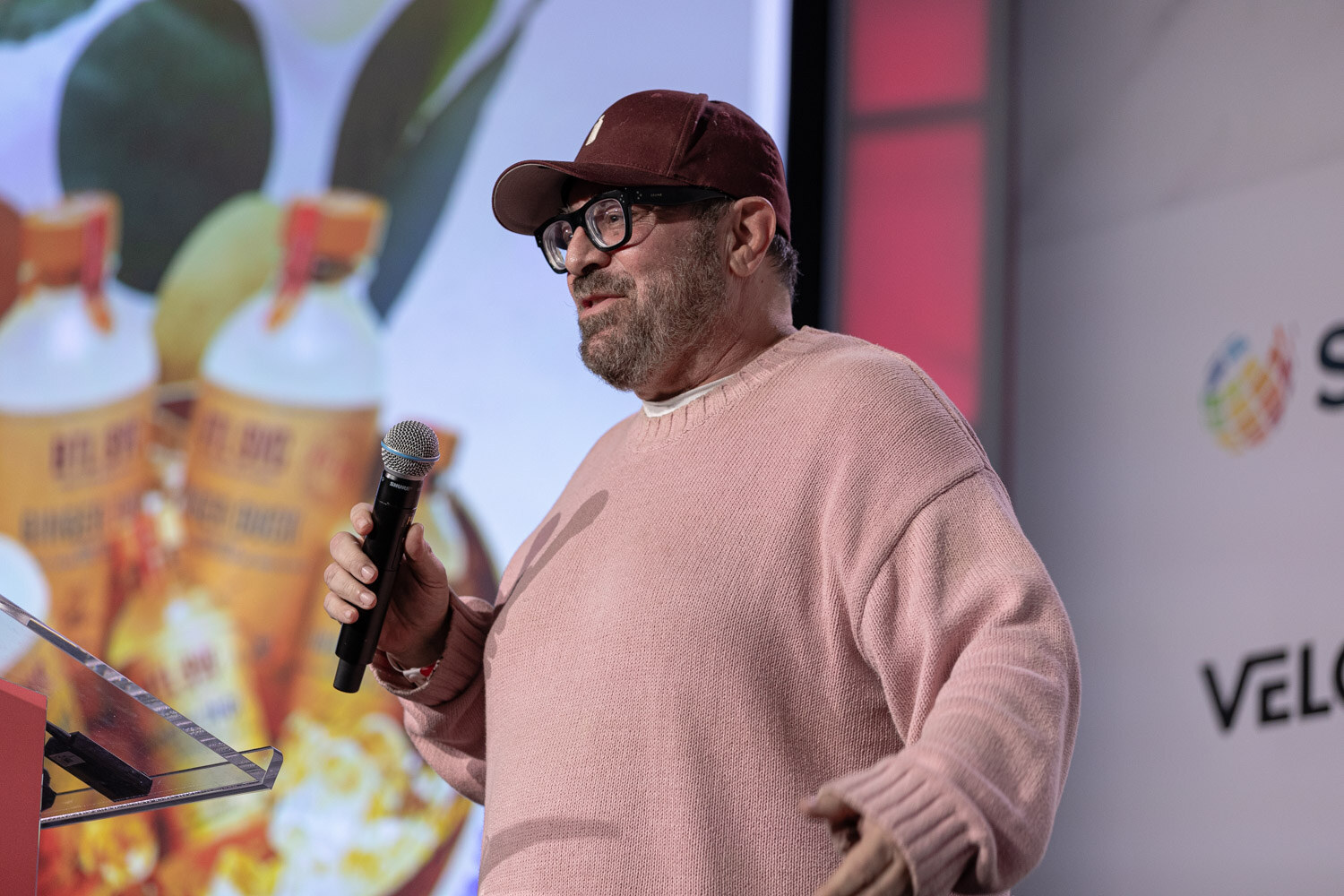 A man in a maroon cap, glasses and pink sweater speaks into a microphone in front of a slide of two bottles.