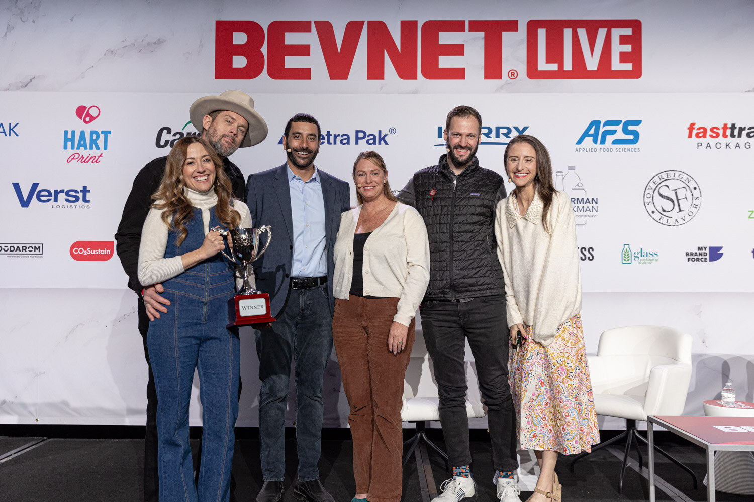 Seven people pose in a row on stage in front of a BevNET Live backdrop, one holding a trophy.