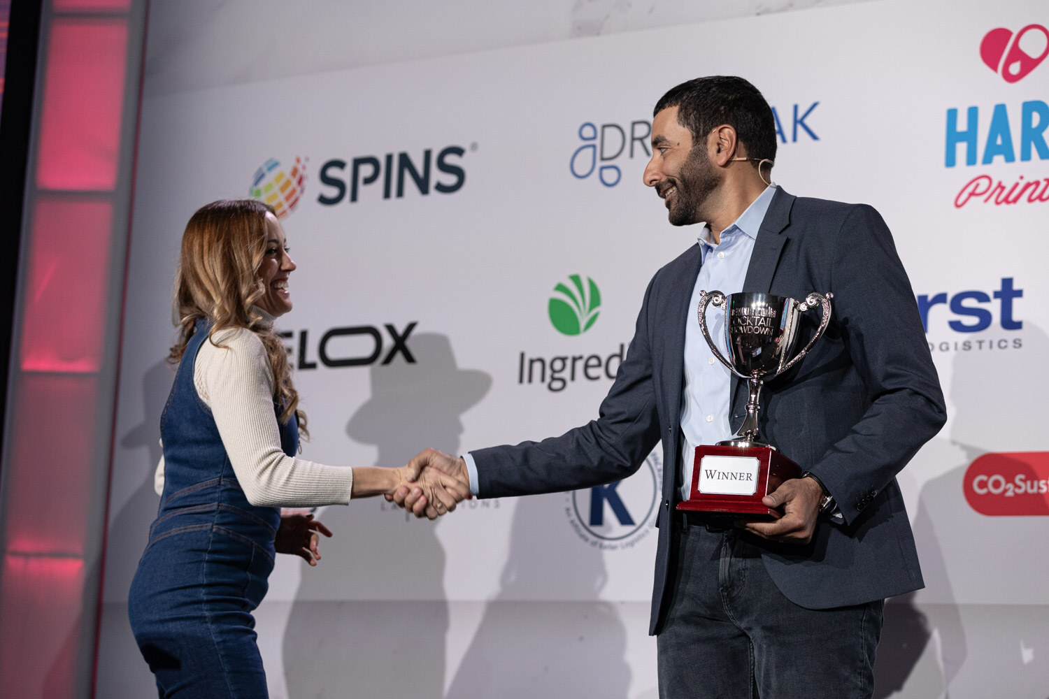 A woman in a denim dress shakes hands with a man in a navy blazer who holds a silver trophy.