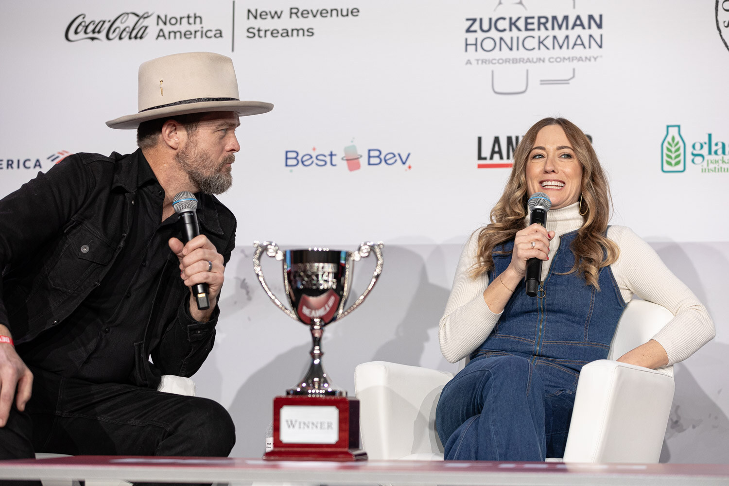 A man in a cowboy hat and a woman in a denim dress sit on stage with a silver trophy between them.