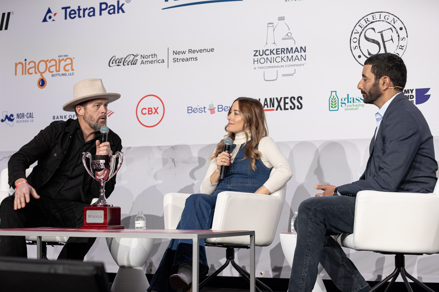 A man in a cowboy hat, a woman in a denim dress and a man in a navy blazer sit on stage with a trophy.