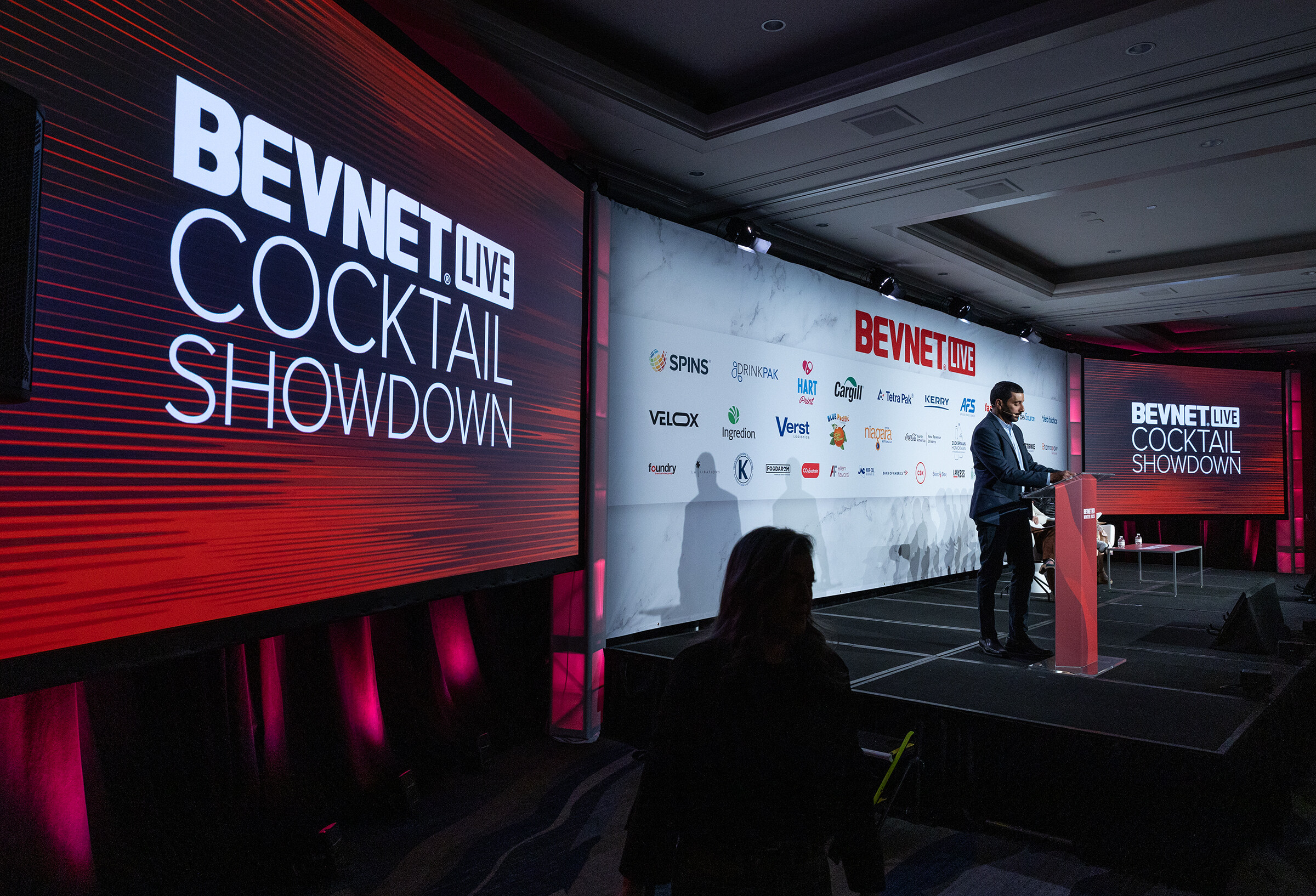 A man stands at a lectern on a dark stage with large red Cocktail Showdown screens.