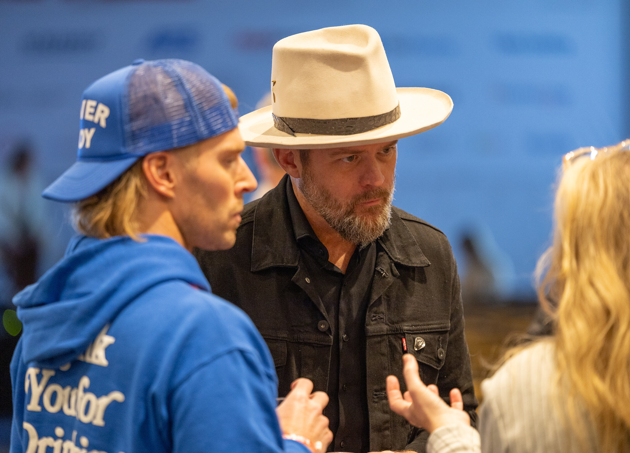 A man in a blue hoodie and a man in a cowboy hat talk in front of a blurred crowd.